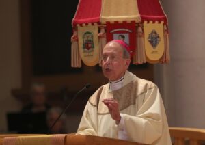 Catholic bishop in cream vestments delivering a sermon at a pulpit, with ceremonial banners in red and gold behind him.