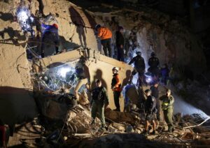 Rescue workers and soldiers on a shattered concrete wall at night, navigating rubble with ropes and flashlights during a search operation.