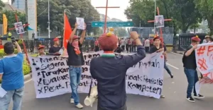 Protest on a city street with several people holding large white banners with handwritten text and red flags; a person at the center faces away, raising a fist.