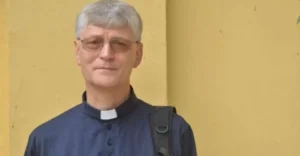 Clergyman wearing a clerical collar and glasses stands outdoors against a yellow wall.