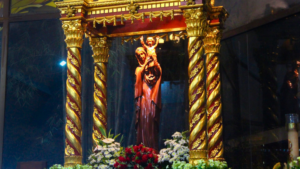 Statue of Mary holding baby Jesus in a gilded, ornate altar with twisted-column pillars and floral arrangements at the base.