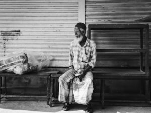 Elderly man with a beard and cap sits on a bench beside a closed storefront, a bagged bundle on a table to his left.