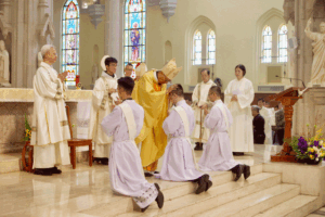 Bishop blessing kneeling altar servers during a Catholic mass inside a church with stained-glass windows.