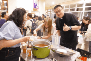 Priest in a black shirt and volunteers ladle soup from a large green pot at a community meal, with people waiting in line behind them.