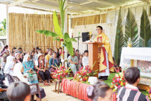 Priest in gold vestments delivering a sermon at a wooden podium during an outdoor religious ceremony, with attendees seated and floral decorations nearby.