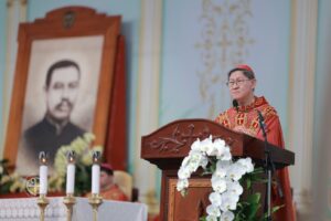 Catholic priest in red vestments delivers a sermon at a carved wooden pulpit adorned with white orchids, microphone nearby.