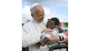 Smiling elderly religious leader in white robes holding a baby outdoors.