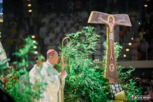 Catholic priest in white vestments holding a ceremonial staff during a service, with a large crucifix and greenery nearby and a blurred crowd in the background.