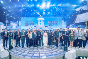 Group of people in formal attire raising glasses in a toast on a circular stage with a blue-lit event set behind them, for a celebratory group photo, wide-angle view.