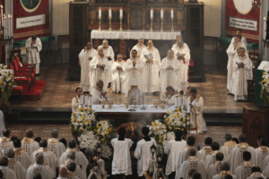 Catholic Mass with many priests around a flower-adorned altar during a liturgy in a church setting