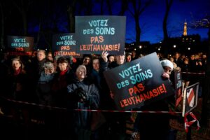 Crowd of protesters at night holding blue and orange signs reading 'Votez des soins, pas l’euthanasie' against euthanasia in a park setting.