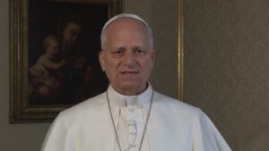 Religious leader in white liturgical robes speaks to the camera indoors, with a framed painting behind.