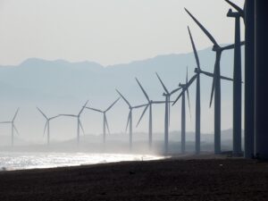 Row of wind turbines along a misty coastline with distant mountains in the background, silhouetted against the sky on the beach edge.