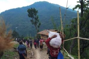 Rural mountain path with villagers carrying large sacks on their shoulders toward a thatched hut, mountains in the background and bamboo fencing along the trail.