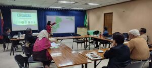 Workshop in session: presenter points to a whiteboard covered with green sticky notes while participants sit around tables in a conference room, projector on screen behind them.