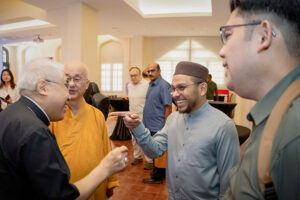 Group of men chatting and laughing at a bright reception hall, a younger man in a traditional cap points as he talks to an older man in orange robes nearby.