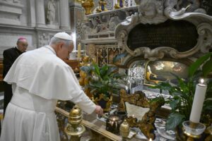 Pope in white robes lighting a candle at an ornate Catholic altar with gold decorations and plants