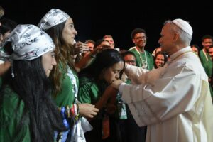 Priest in white robes blessing a young woman who kisses his hand, surrounded by smiling peers in green shirts and lanyards at night.