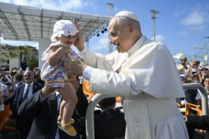 Pope in white robes blessing a baby held up by a man at a sunny outdoor event, with a cheering crowd in the background.