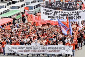 Large crowd of protesters marching on a street with banners and flags, condemning political issues in Filipino/Tagalog.