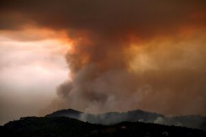 Massive smoke plume and orange glow from a wildfire over a forested hillside at dusk, with low hills in the foreground in shadow.