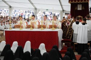 Priests in white and gold vestments at an ornate altar under a white tent, with a bishop in pink hat and a crowd in the background; nuns in the foreground observe.