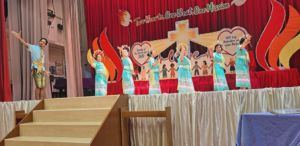 Group of dancers in turquoise traditional outfits perform on a stage with a red curtain backdrop and a banner that reads 'Two hearts, One Beat, One Mission'.