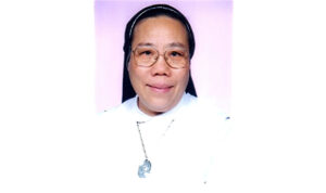 Portrait of a smiling nun wearing glasses and a white habit against a pink background.”,