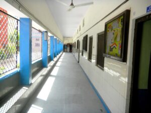 Long indoor hallway with blue support columns, metal window grilles on the left, and framed posters along the right wall.