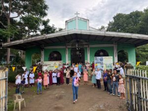 Group of people gathered outside a green church for a community event, with banners and a cross on the roof.