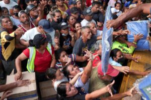 Crowd of people tightly packed at a relief distribution, reaching for bags and boxes being handed out. Pinched expressions show urgency and chaos in the queue.