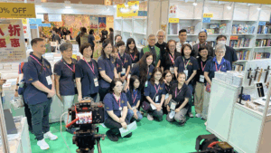 Group of event staff and attendees posing for a photo at a bookstore exhibition booth, with shelves of books and a camera on a tripod in the foreground.