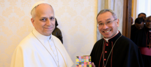 Two smiling clergymen, one in white papal vestments and the other in black clerical robes, pose together and hold a colorful patterned book or card between them.