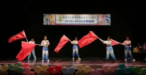 Five dancers on a dark stage wave large red flags; balloons line the front and a banner reading 2025-2026 hangs overhead.