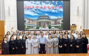 Group of nuns and attendees posing for a formal group photo on a stage in a hall, with a large screen showing a domed campus building and a red banner in the background.