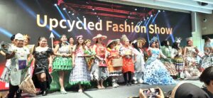 Group of models in colorful upcycled outfits posing on stage at a show backdrop reading 'Upcycled Fashion Show'.