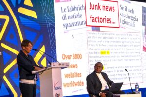 Woman at a podium presenting, with a large screen behind showing the headline 'Junk news factories...' and a man with a laptop seated nearby.