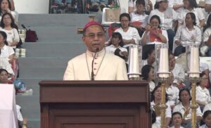 Catholic bishop in cream vestments and pink skullcap delivers a sermon from a wooden pulpit, with a crowd in white shirts seated behind and tall candle stands nearby.