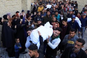 Crowd of mourners carries a wrapped body through a street, many raising hands in grief.