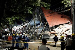 Emergency responders and bystanders search through rubble after a collapsed building at night, with caution tape and debris visible in the scene.
