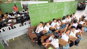 Group of young students in white shirts and plaid skirts sit at wooden desks in a classroom with large green chalkboards displaying writing in the background.