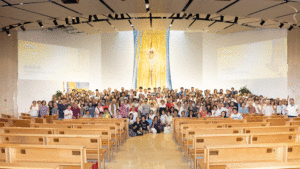 Large group portrait in a church-like auditorium with wooden pews and a gold/blue tapestry behind a central statue on stage.