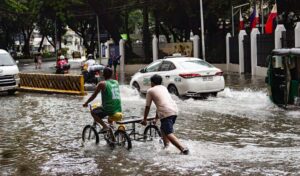 Two men navigate a flooded city street: one on a bicycle with a cart, the other pushing the cart through waist‑deep water as cars pass by.