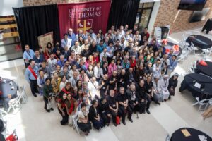 Large diverse group of people posing for a group photo in a campus atrium with a University of St. Thomas banner.