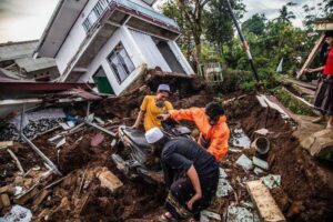 Rescuers move a motorcycle through mud and debris amid a collapsed home after a disaster.