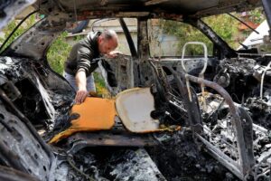 Man inside a severely burned-out car, leaning over to inspect the charred interior amid ash and melted metal.