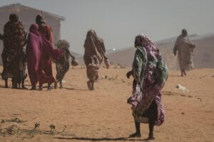 People in colorful robes walk across a sandy desert toward a camp with tents in the background.