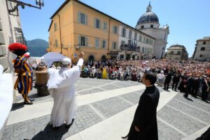 Cleric in white robes raises arms to bless a crowded town square in Italy, with a colorful performer on the left and a domed church in the background.