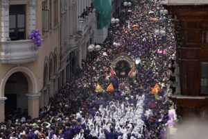 A crowded city street parade with hundreds of people wearing purple and white, carrying a ornate religious float centered among the crowd.