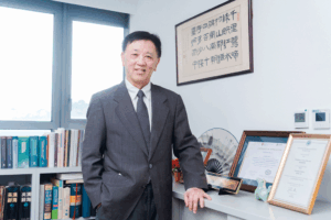 Smiling businessman in a gray suit stands beside a bookshelf and framed certificates in an office setting.